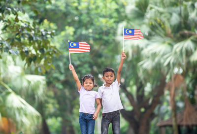 Portrait of siblings holding malaysian flag while standing against trees at park