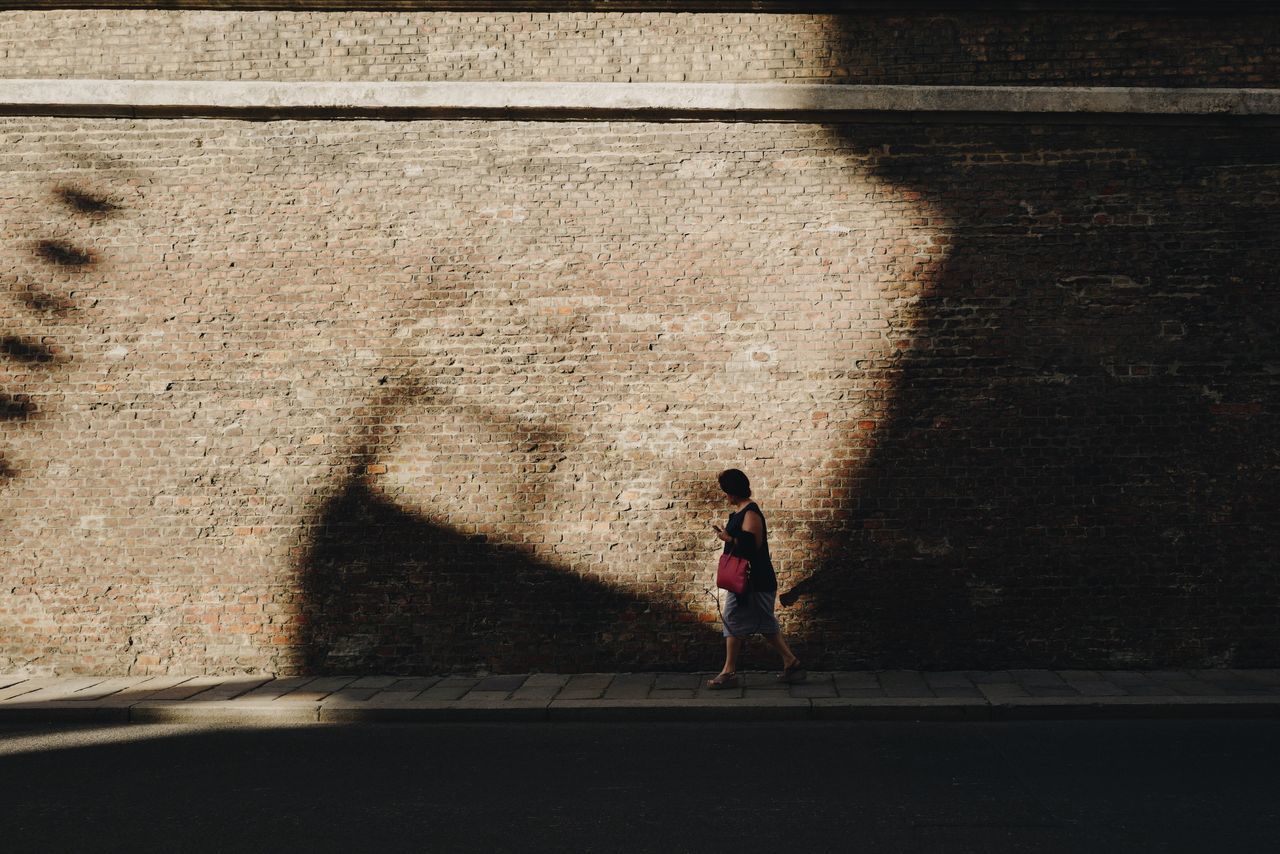 full length, shadow, one person, sunlight, real people, walking, architecture, wall - building feature, women, footpath, day, lifestyles, built structure, child, nature, wall, adult, sidewalk, city, outdoors