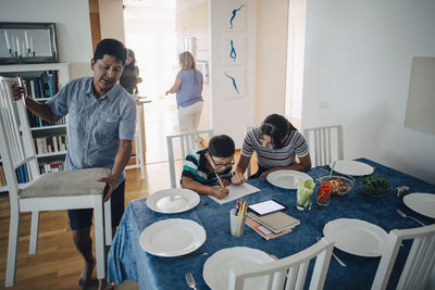 Teenage girl assisting brother in homework while father arranging chairs at dining table