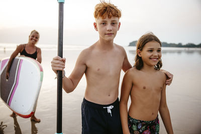 Portrait of shirtless boy with arm around sister at beach during vacation