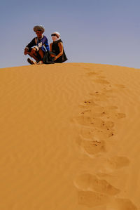 People sitting on sand dune in desert against clear sky