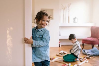Smiling girl standing by wall while brother playing in background at home