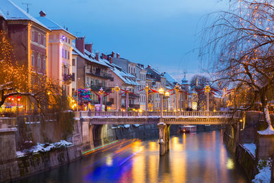 Illuminated bridge over river by buildings against sky at dusk