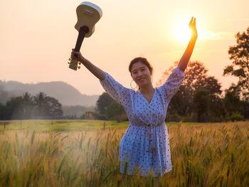 Portrait of young woman with arms raised standing on field against sky