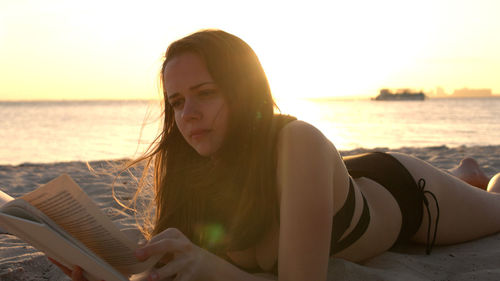 Portrait of young woman sitting at beach against sky during sunset