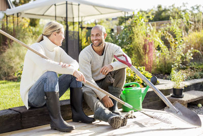 Full length of mature couple with gardening equipment sitting at yard