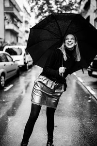 Cheerful woman holding umbrella while standing on road in city