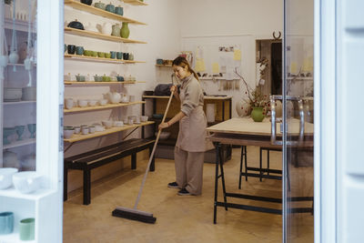 Female potter cleaning floor with broom while standing at workshop