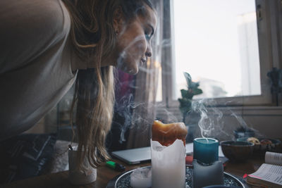 Side view of young woman blowing scented candles on table against window in dorm room