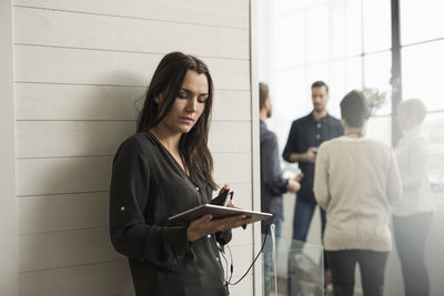 Woman holding digital tablet while leaning on wall with people in background