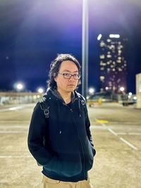 Portrait of young asian man standing in deserted rooftop parking lot against buildings at night