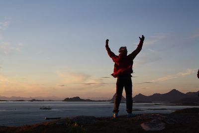 Man with arms raised standing on cliff against sea during sunset