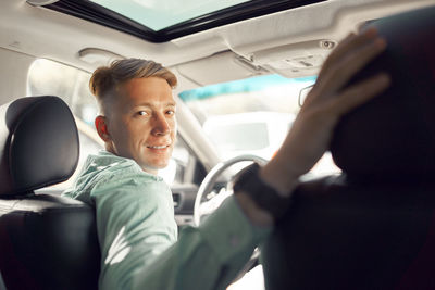 Side view of handsome smiling male driver sitting in parked luxury car and looking at camera