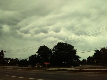 Silhouette trees on country road against cloudy sky