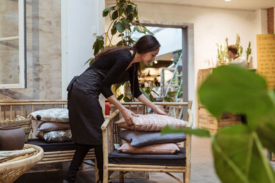 Young saleswoman stacking cushions on chair at store