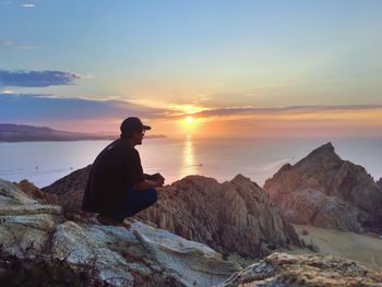 Man sitting on rock at sea shore against sky during sunset