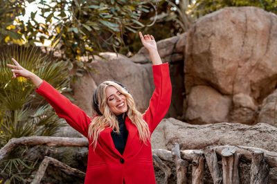 Portrait of smiling young woman with arms raised standing against rock