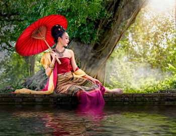 Young woman with umbrella sitting by lake in forest