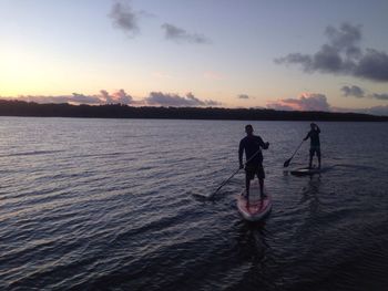 Silhouette people paddleboarding in river against sky at dusk