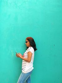 Young woman using phone while standing against blue wall