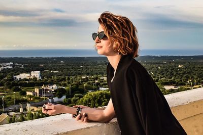 Side view of young woman looking at cityscape against sky