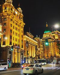 Illuminated city street by buildings against sky at night