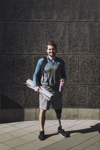 Portrait of smiling man with disability holding water bottle and exercise mat on footpath during sunny day