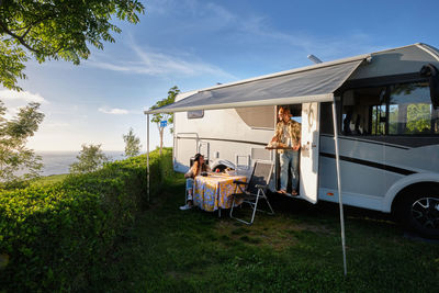 Cheerful traveling couple of hipsters having dinner at table near caravan during trip in summer in nature