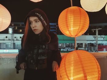 Woman looking away while standing by illumianted lanterns at night