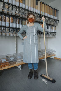 Female store clerk with hand on hip holding broom while standing in zero waste shop during covid-19