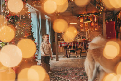 Candid authentic happy little boy in knitted beige sweater hugs dog with bow tie at home on xmas