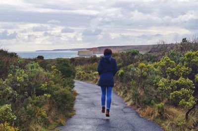 Rear view of woman walking on road leading towards sea against cloudy sky