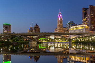 Illuminated bridge over river by buildings against sky at night