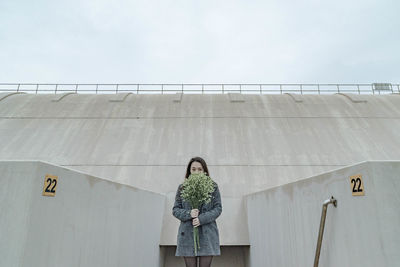 Young woman with flower bouquet standing against surrounding wall