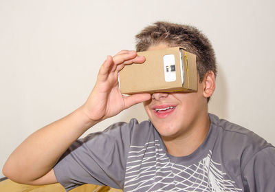 Smiling teenager boy covering eyes with smart phone in cardboard box