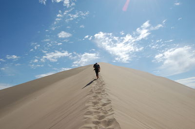 Rear view of man climbing on sand dune at desert against sky during sunny day