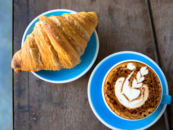 Cup of hot cappucino in blue cup with croissant on wooden table background