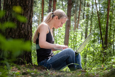 Girl in the forest sits leaning against a tree, working on a laptop. digital detox. mental health