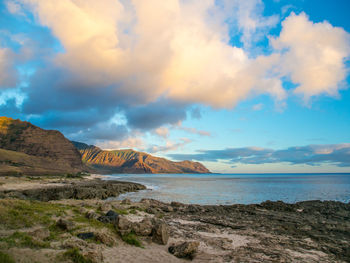 Scenic view of sea by rocky mountains against cloudy sky during sunset at oahu