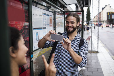 Smiling man photographing female friend through smart phone at bus stop in city