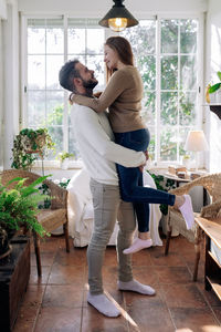 Smiling woman riding piggyback on boyfriend while looking at each other against windows at home