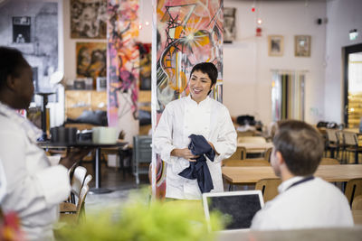 Smiling female chef holding apron discussing with multiracial colleagues in restaurant