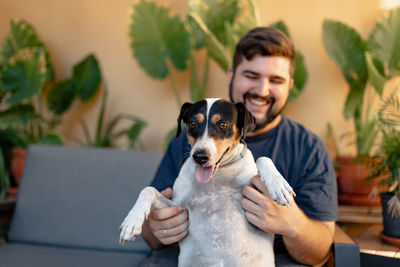 Young man smiling is holding his dog while playing with his paws. the dog is looking at camera