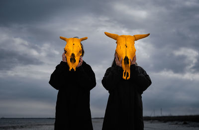 People with yellow mask standing by sea against cloudy sky