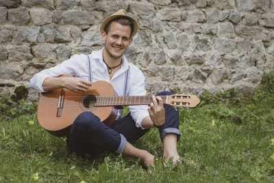 Portrait of young man playing guitar while sitting on grass against wall