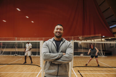 Smiling male coach standing with arms crossed at badminton court