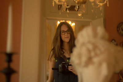 Beautiful young woman holding camera while standing at home