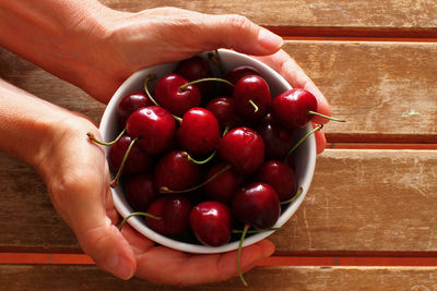 Overhead photo of hands holding up a white bowl with cherries on wooden slatted table.