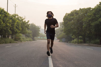Front view of a man wearing mask jogging on an empty road during sunrise.