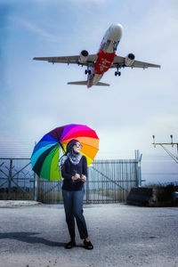 Full length of woman standing while holding umbrella against airplane in sky